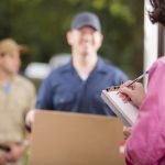 Home delivery. Men deliver large package to customer at her front door. She signs her name to delivery confirmation form on clipboard.  Mail order.  Men wear uniforms. Large cardboard box.