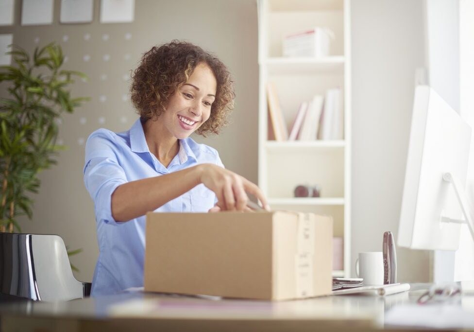 a young woman in her home office looks happy as she unpacks a small cardboard box containing something exciting